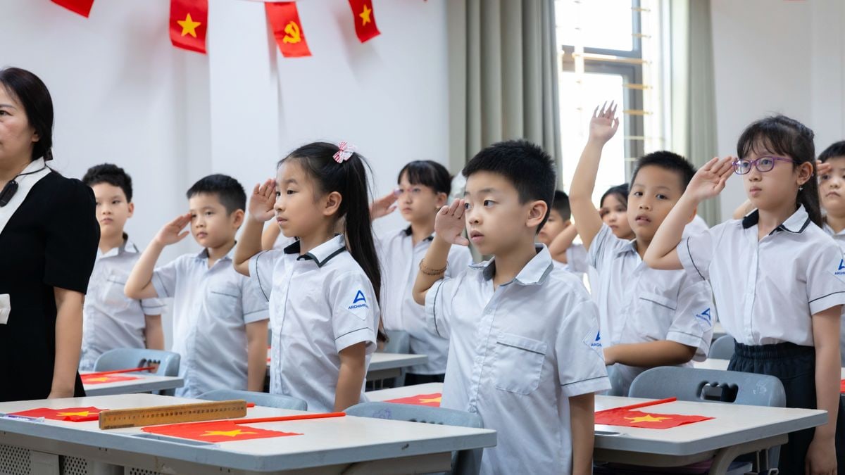 Vietnamese students in white uniforms salute flags during a classroom activity. Bright indoor setting.